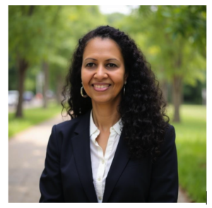 Portrait of a woman with loose curly hair and olive skin tone wearing a dark blazer and white button‑down shirt, standing on a tree‑lined path outdoors, facing the camera with a slight smile.